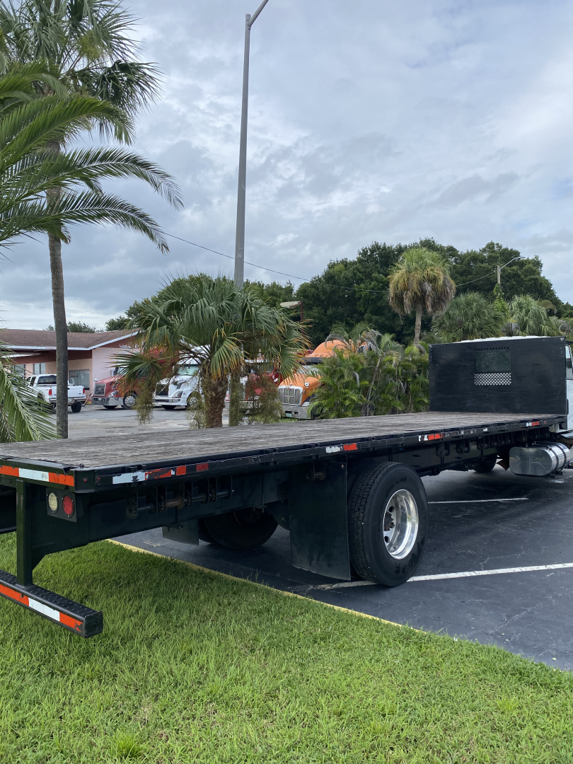 Flatbed tow truck chassis with long black flatbed and rear wheel, cabless area at the front, some palm trees and vehicles in background.