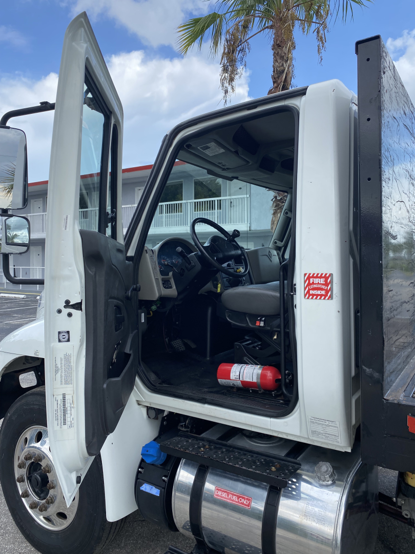 Cab of a white 2018 International 4300 truck with door open, showing steering wheel, dashboard, and cylindrical diesel tank under door; red fire extinguisher mounted inside.