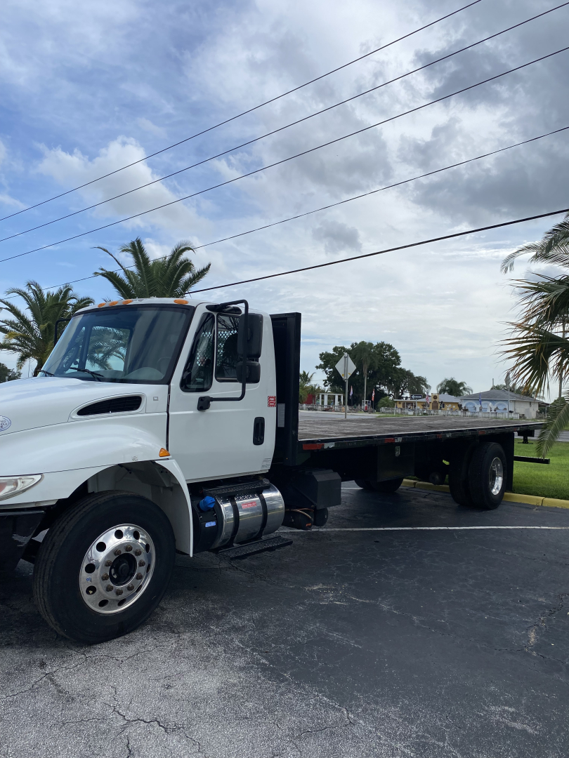 2018 International 4300 flatbed truck in a parking lot, front left cab visible with grille, headlights, and side mirror; long flatbed with dual rear wheels.