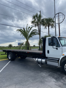 Flatbed tow truck with white cab and empty black trailer showing exterior details and palm trees in the background.