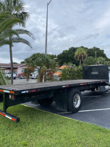Flatbed tow truck chassis with long black flatbed and rear wheel, cabless area at the front, some palm trees and vehicles in background.
