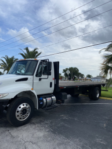 2018 International 4300 flatbed truck in a parking lot, front left cab visible with grille, headlights, and side mirror; long flatbed with dual rear wheels.