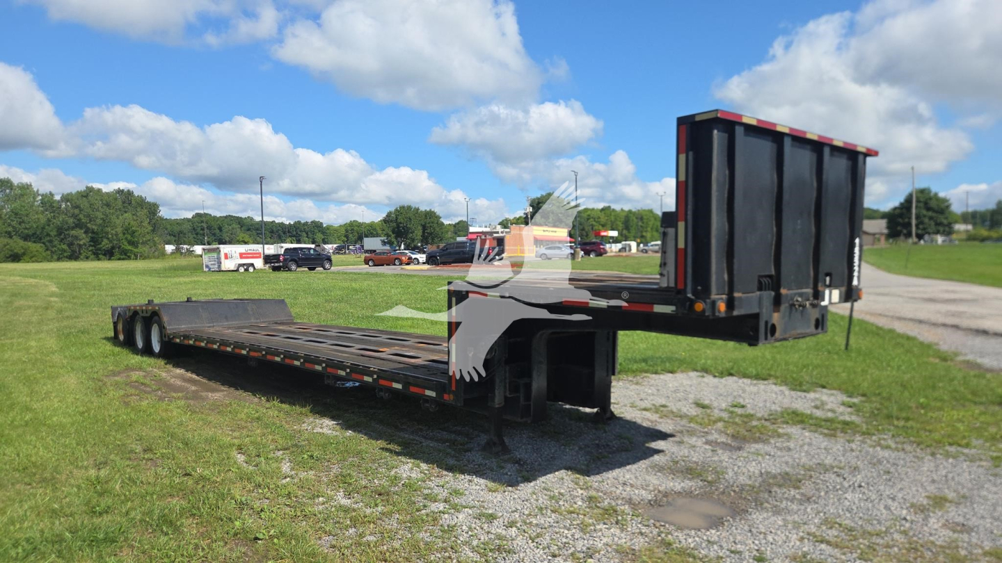 Lowboy trailer with gooseneck and folded rear ramp in field; black trailer deck with metal supports and white/gray accents; clear sky
