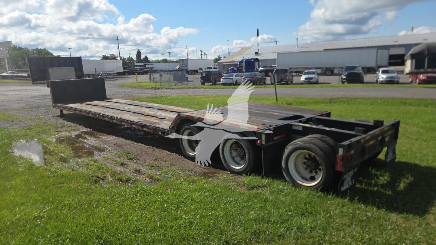 Lowboy trailer with long deck and dual rear axles, side rails, and flatbed surface visible; VIN/stock not legible; yard with buildings and vehicles in background.