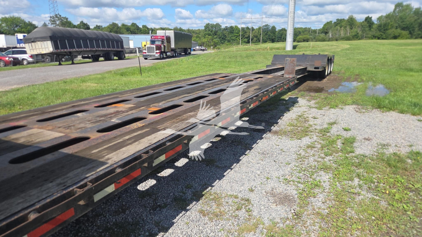 Lowboy trailer with long steel deck and gooseneck, viewed from side showing multiple cutouts and deck slots; open grass field and other trailers in background.