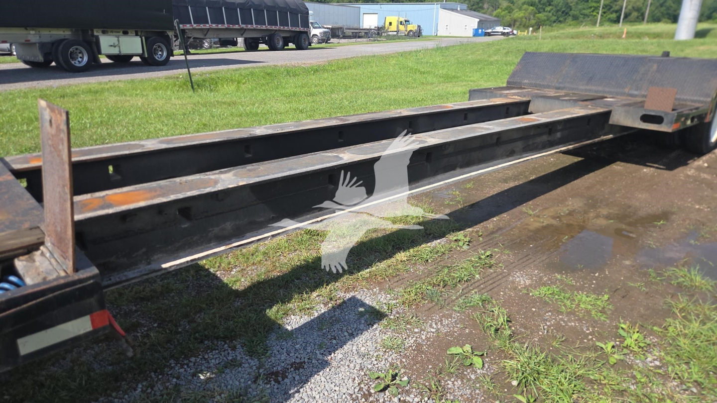 Trailer frame showing longitudinal steel beams and deck of a Trail King TK90ED482 lowboy trailer, with visible rusted edges and grass around.