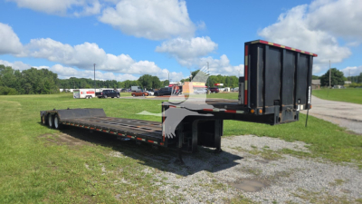 Lowboy trailer with gooseneck and folded rear ramp in field; black trailer deck with metal supports and white/gray accents; clear sky