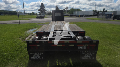 Rear view of a flatbed trailer showing taillights, reflectors, and dual rear axles. Visible text includes EAST MFG CO, RANDOLPH, IA on the bumper and license-like plate area.