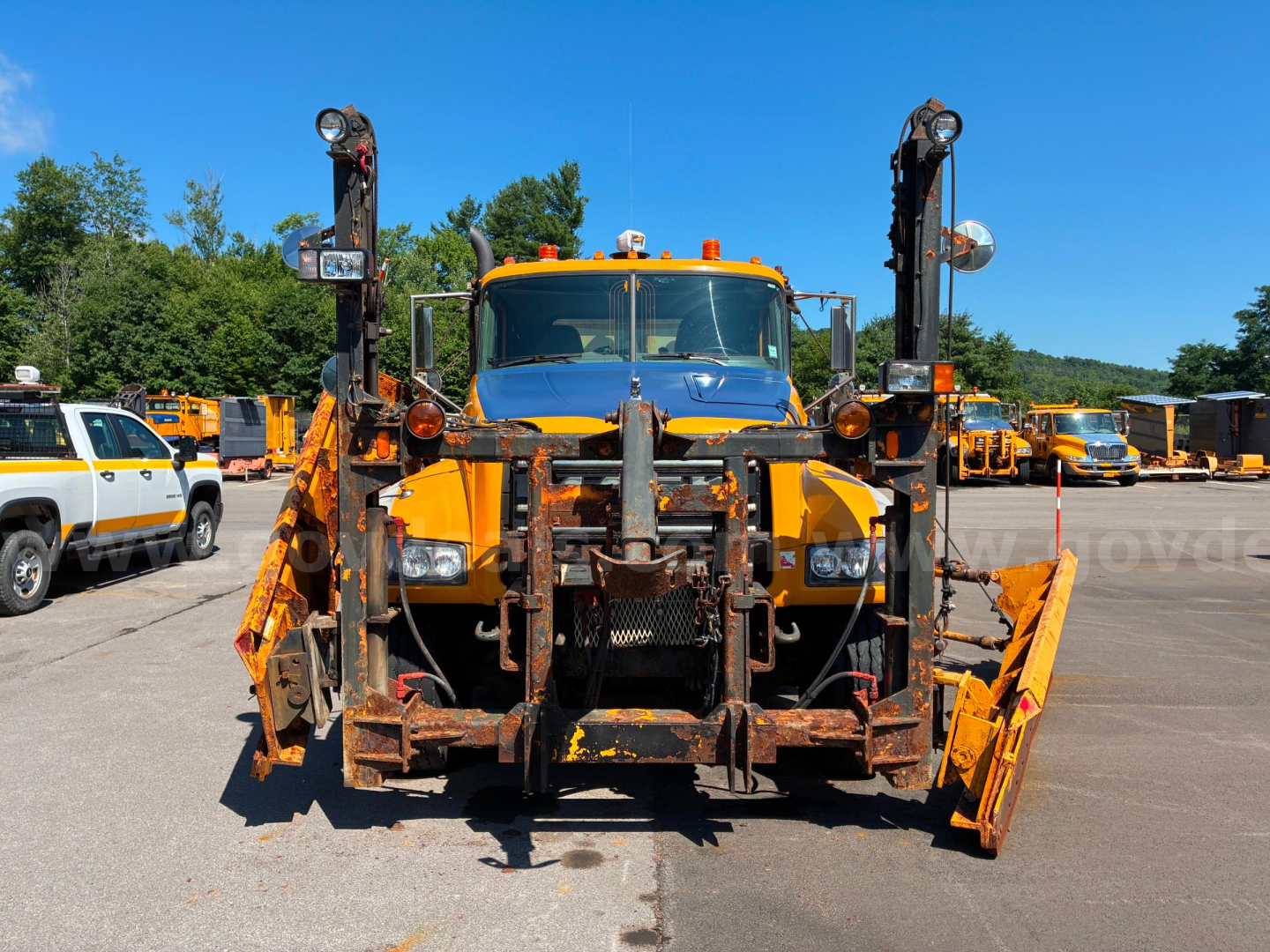 Front view of a yellow 2013 Mack GU713 diamond-tump truck with snowplow and front-end equipment in a lot.