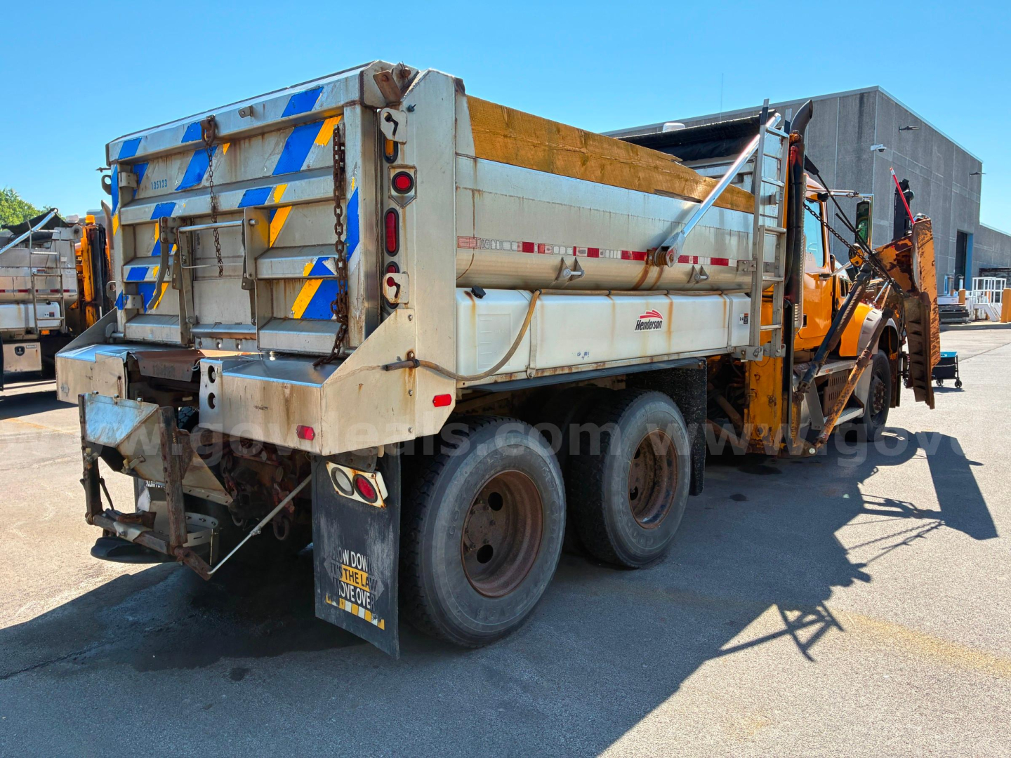 Mack GU713 dump truck rear-side view showing hydraulic bed, dual rear axles, and tailgate with blue/yellow reflective stripes.