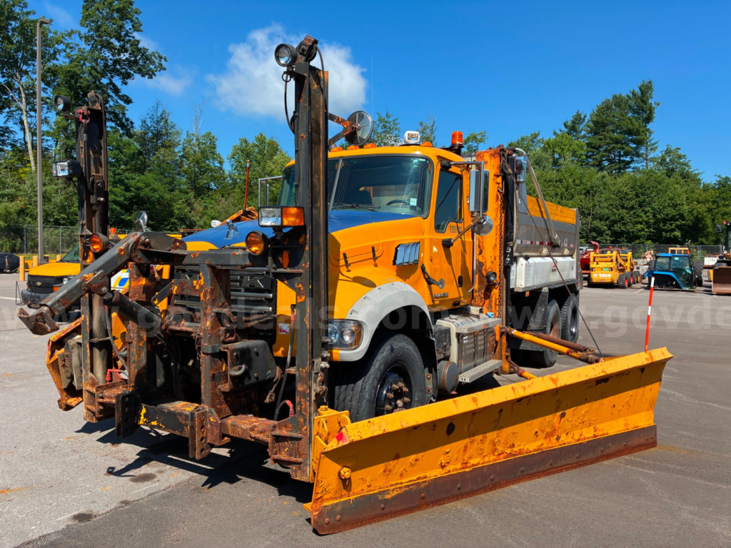 2013 Mack GU713 dump truck in-service showing snow plow attachment and hydraulic equipment, orange cab with dump bed, large front bumper, and black tires.