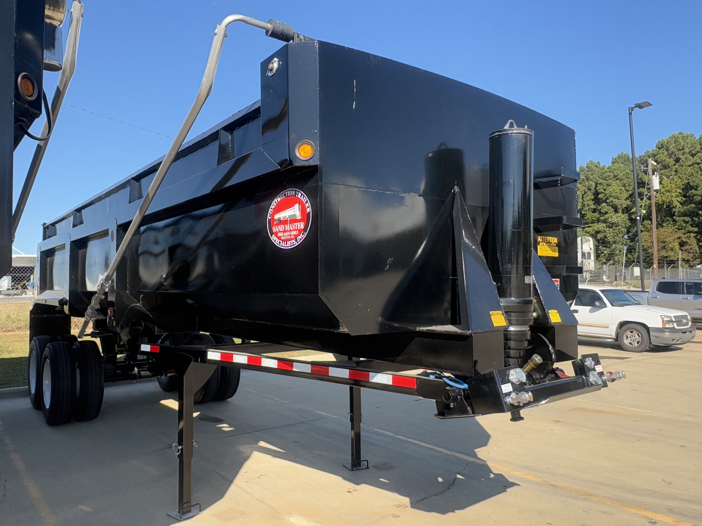 Dump trailer showing black steel dump body with side railing and front hydraulic cylinder, stabilized by landing gear, parked in lot.