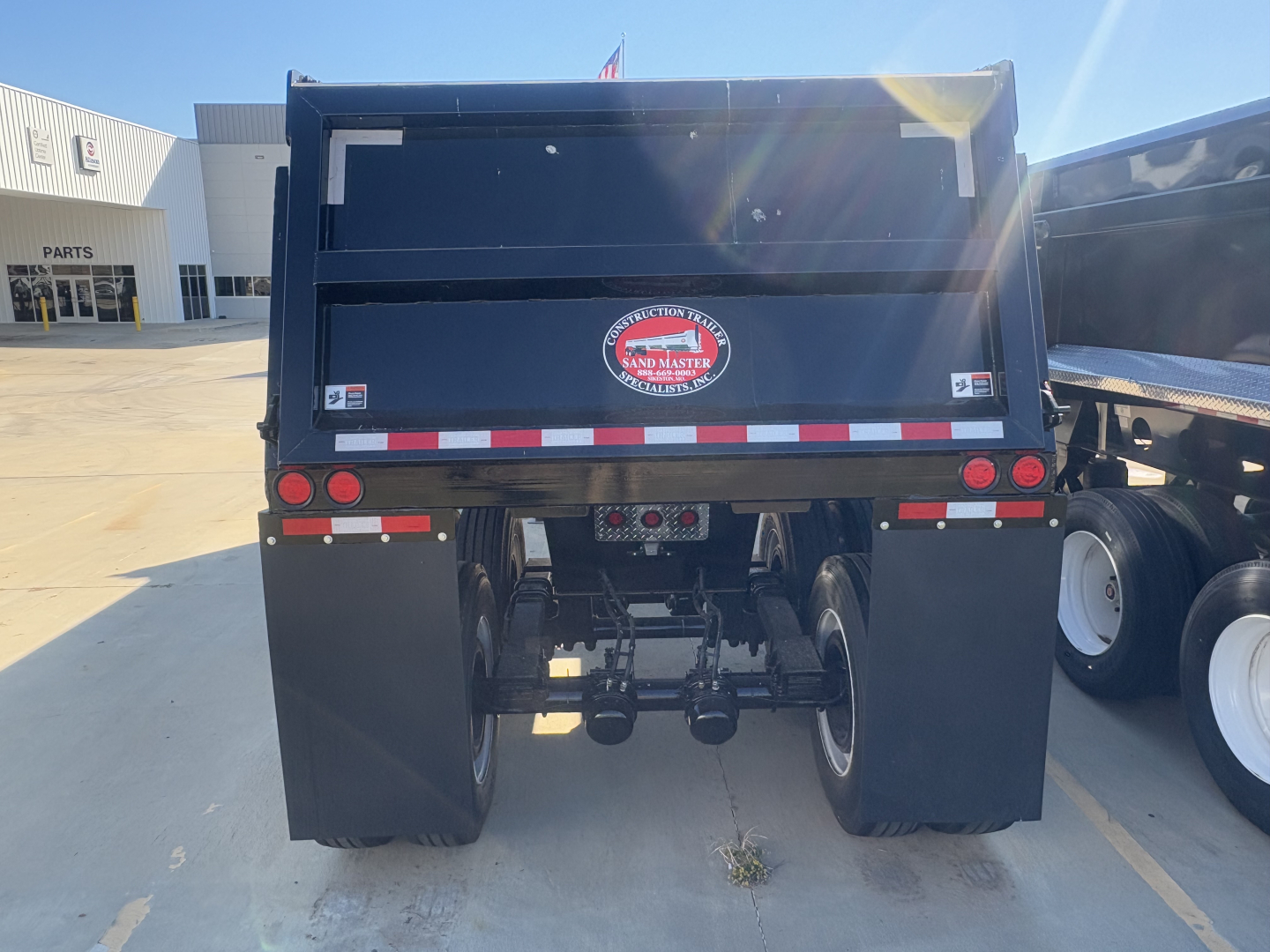 Dump trailer rear view showing mud flaps, taillights, and hitch assembly with Sand Master logo plate; reflective white/red stripes along rear edge; black steel structure.