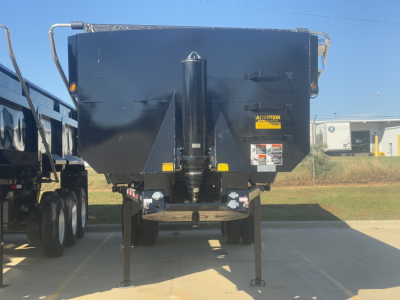 Dump trailer showing rear view with two stabilizer legs, central hitch, and warning label on right side; license plate area not visible; yellow caution sticker near top center.