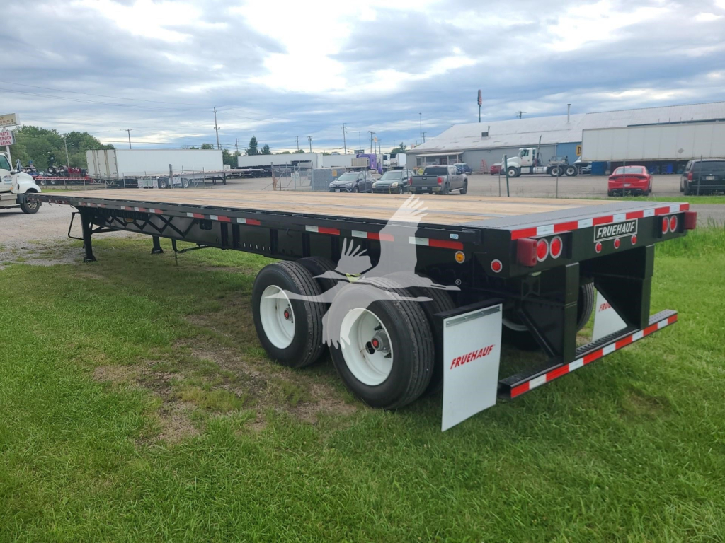 Flatbed trailer showing dual rear axles with white wheels, black chassis, wooden deck, and Fruehauf branding on rear and mud flaps.