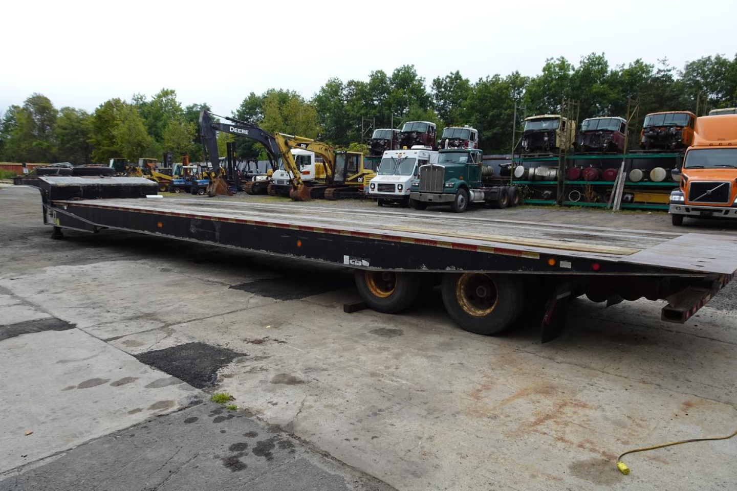 Flatbed trailer in a yard with stacked equipment and trucks in background; long low-profile deck, dual rear axles, worn surface.