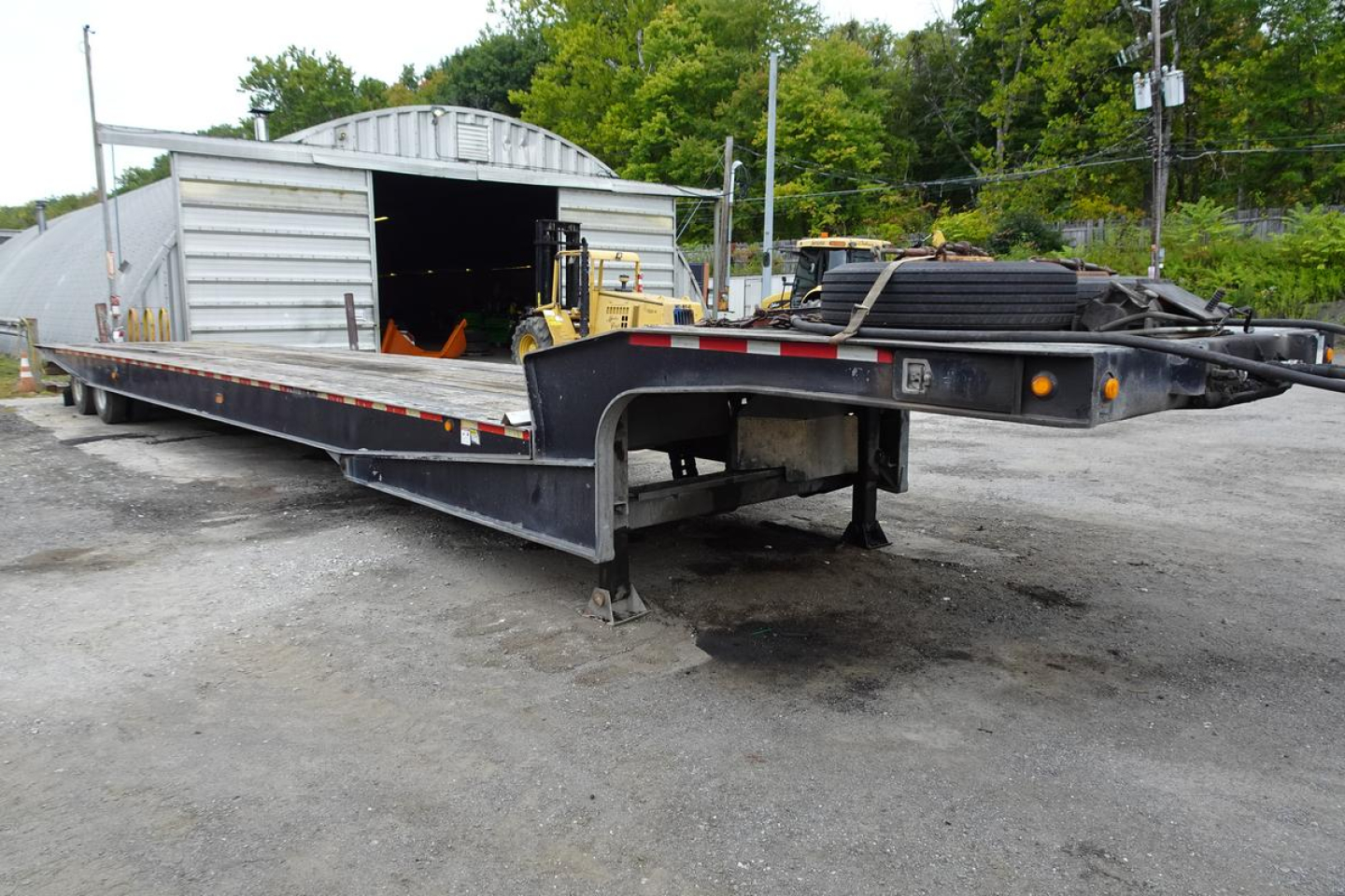 Long flatbed trailer with a large barn-style storage building in background. Trailer shows gooseneck hitch and dual axles, with various equipment and tires on the bed.