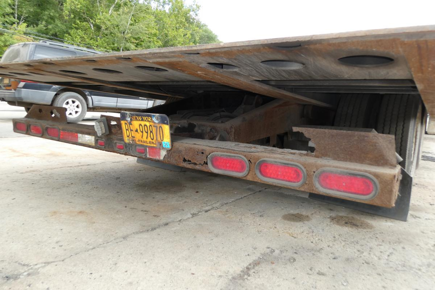 Trailer rear view showing rusted steel landing gear and license plate with New York BE-99870. Red reflective taillights and mud flaps under a flatbed trailer.