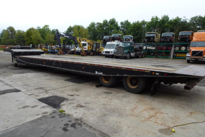 Flatbed trailer in a yard with stacked equipment and trucks in background; long low-profile deck, dual rear axles, worn surface.