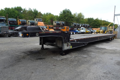 Long lowboy trailer with black frame and orange accents, showing flatbed surface and gooseneck area. Background: multiple trucks and equipment in a lot.