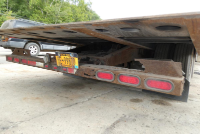 Trailer rear view showing rusted steel landing gear and license plate with New York BE-99870. Red reflective taillights and mud flaps under a flatbed trailer.