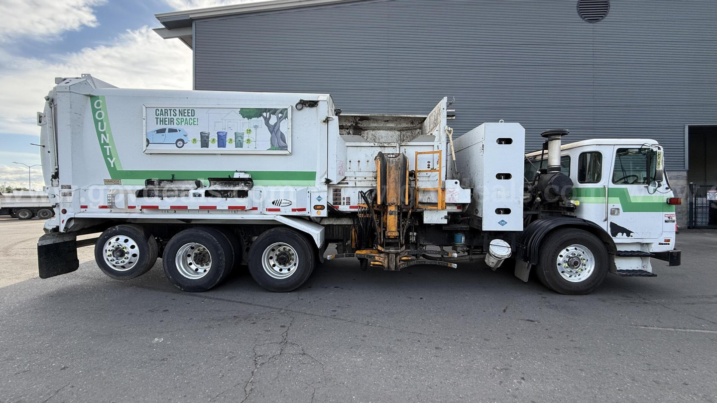 Truck side view of a white Autocar XPeditor garbage truck with multi-axle rear, loading mechanism, and graphic panel on the side.