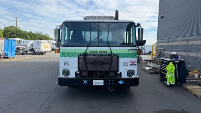 Front view of a white Autocar Xpeditor truck with a large grille, black bumper, and blue-green accents. License plate visible, headlights, mirrors, and emblem on grille.