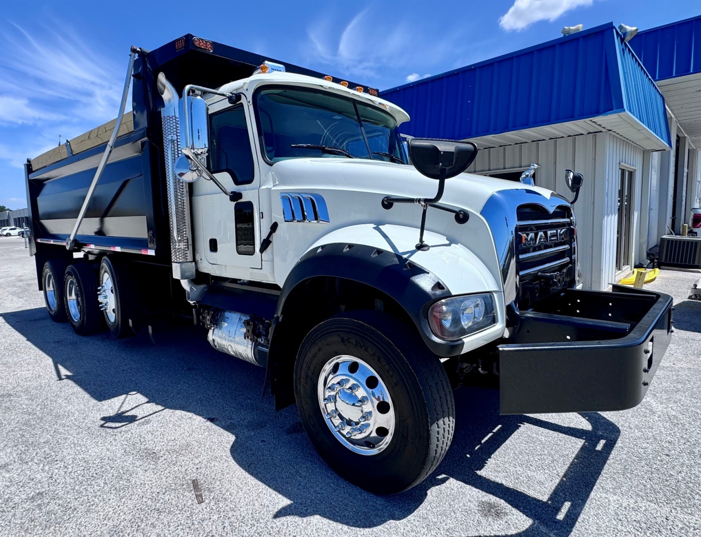Dump truck cab and body of a white 2023 Mack GR84F with chrome wheels and a front bumper mounted toolbox, side-view showing dump bed and blue-building background.