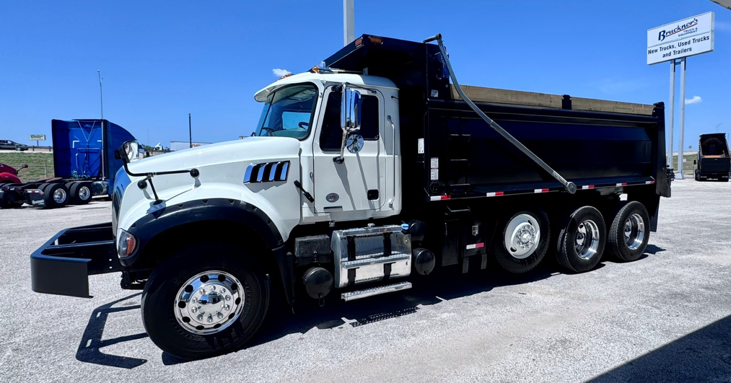 Truck shows a white Mack GR84F dump truck with a black dump bed, side steps, multiple wheels, and front bumper in a gravel lot.