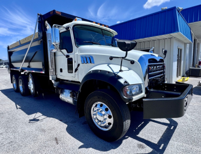 Dump truck cab and body of a white 2023 Mack GR84F with chrome wheels and a front bumper mounted toolbox, side-view showing dump bed and blue-building background.
