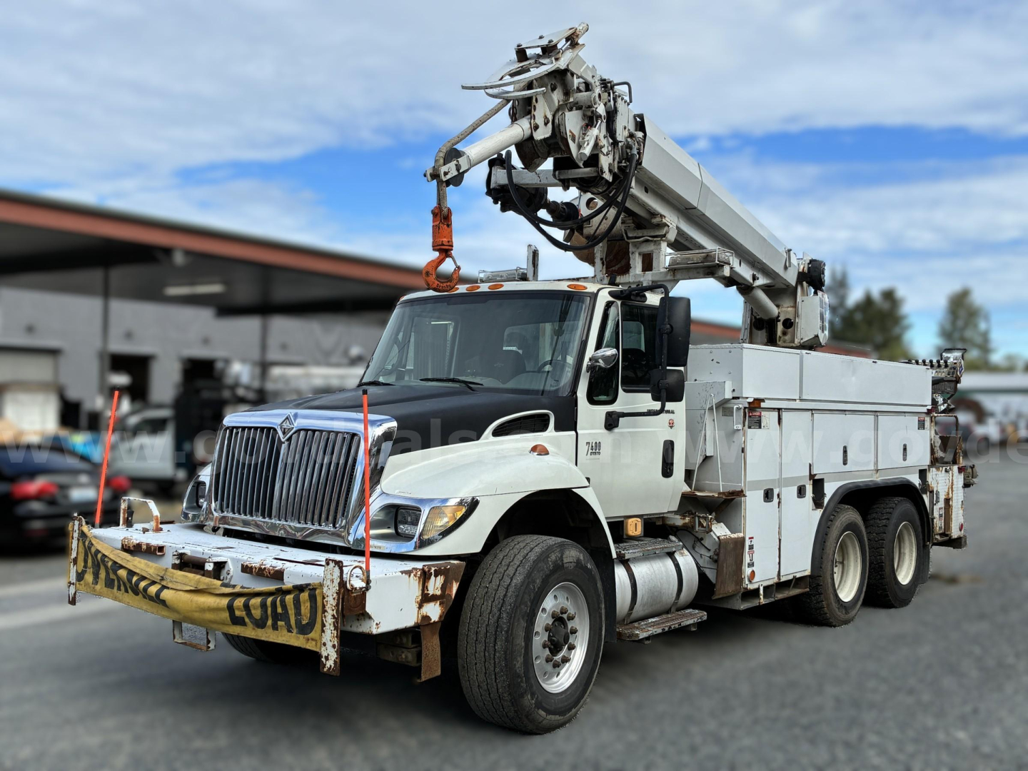International 7400 night crane truck with mounted boom in a work yard, utility body, front bumper with safety rails and visible rust patches.