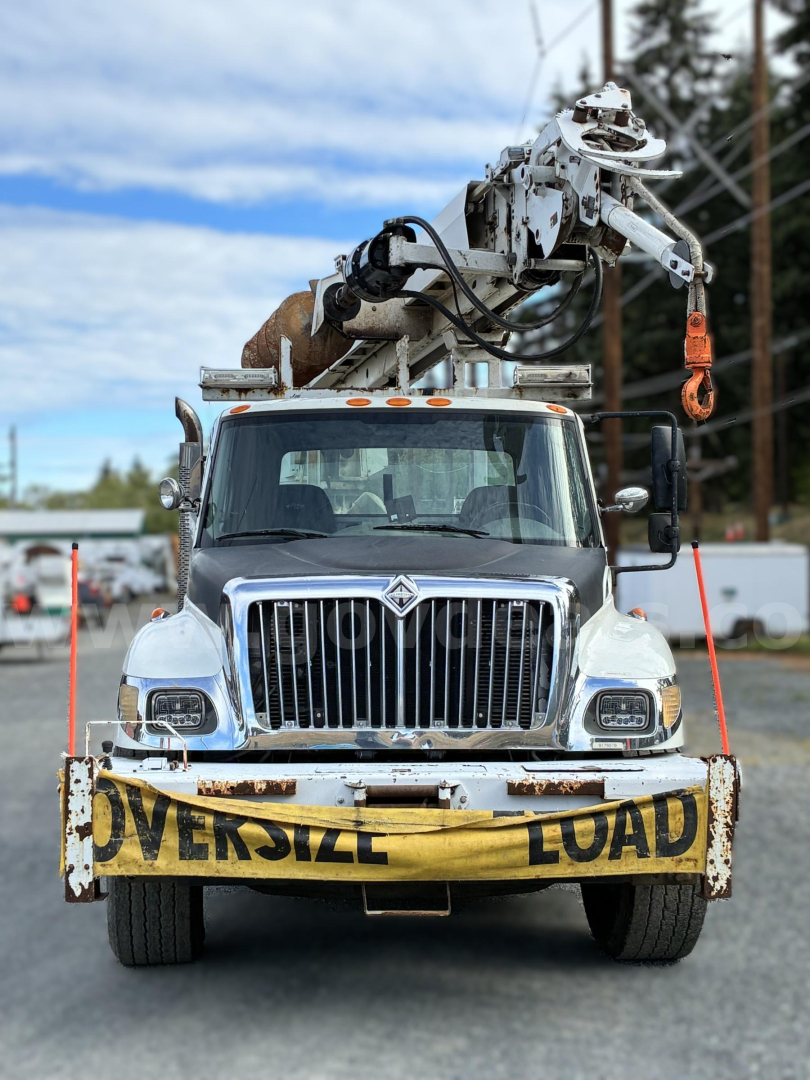 Front view of a 2007 International 7400 crane truck with a mounted derrick crane and orange hook, oversized load banner on bumper, chrome grille, blue cab.