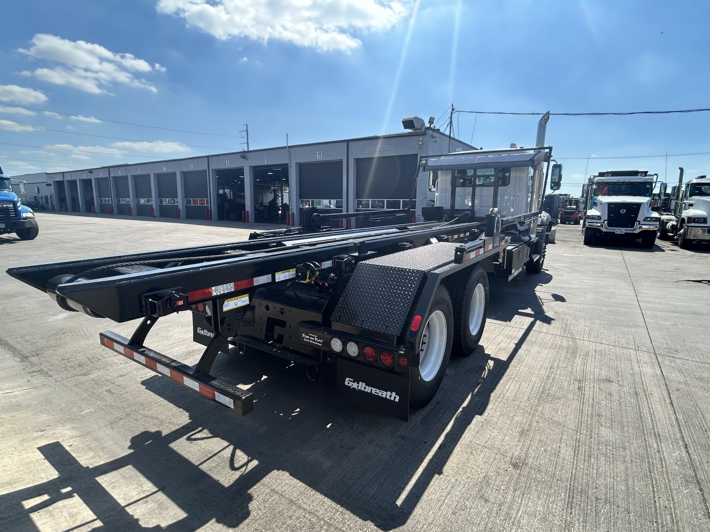 Truck flatbed semitrailer in a lot with a Mack GR64B roll-off chassis and black steel platform.