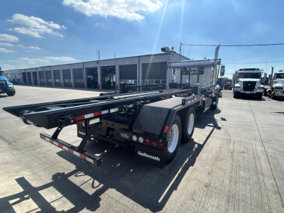 Truck flatbed se­mitrailer in a lot with a Mack GR64B roll-off chassis and black steel platform.