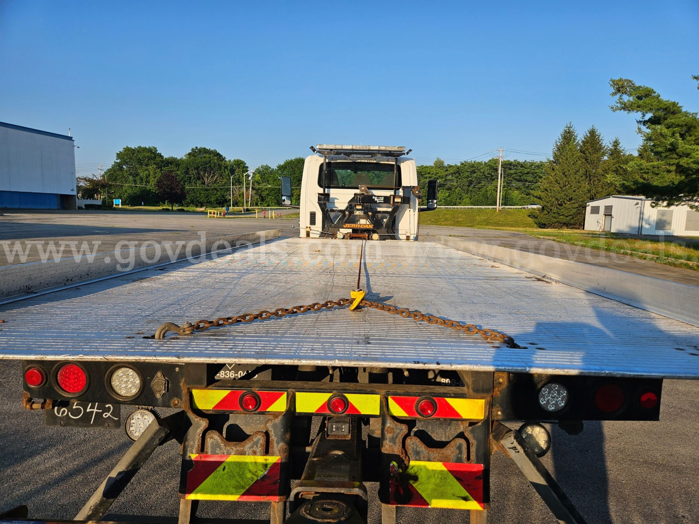 Tow truck flatbed with metal deck and yellow/red reflective safety markings back view, tow chain hanging across bed, winch at cab, clear blue sky.