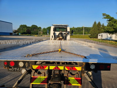 Tow truck flatbed with metal deck and yellow/red reflective safety markings back view, tow chain hanging across bed, winch at cab, clear blue sky.