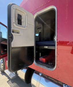 Peterbilt 579 truck sleeper cab side door open showing interior storage and steps, red exterior with black lower panel and chrome fuel tank.