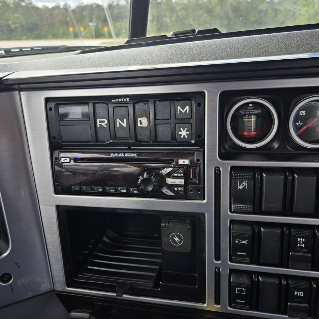 Dashboard cockpit of a 2019 Mack truck showing gear selector panel with R N D M indicators and Mack branded radio, gauges, and switch panel.