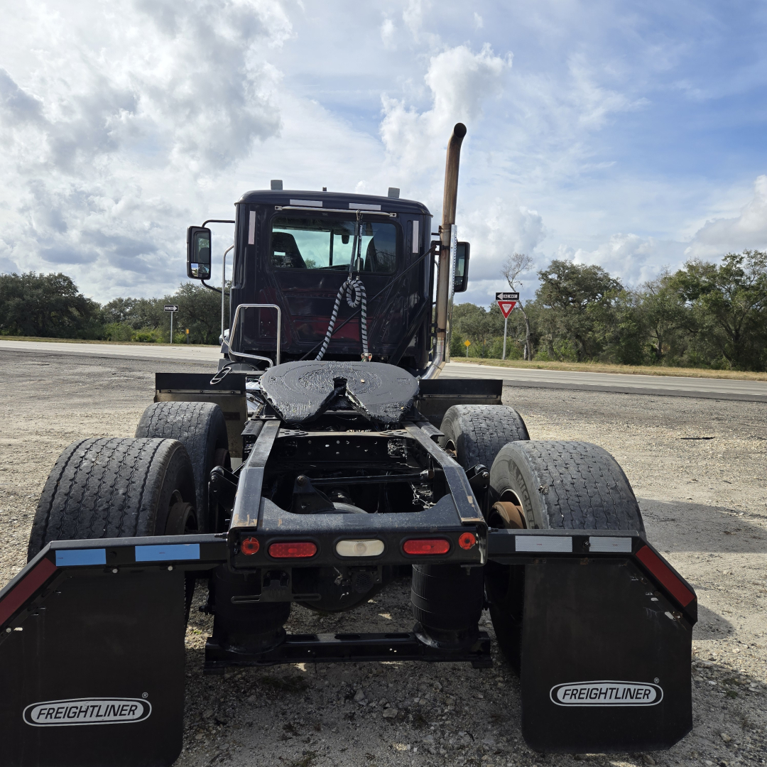 Truck chassis rear view showing dual rear axles, mudflaps with Freightliner logo, fifth wheel, and exhaust stack behind cab.