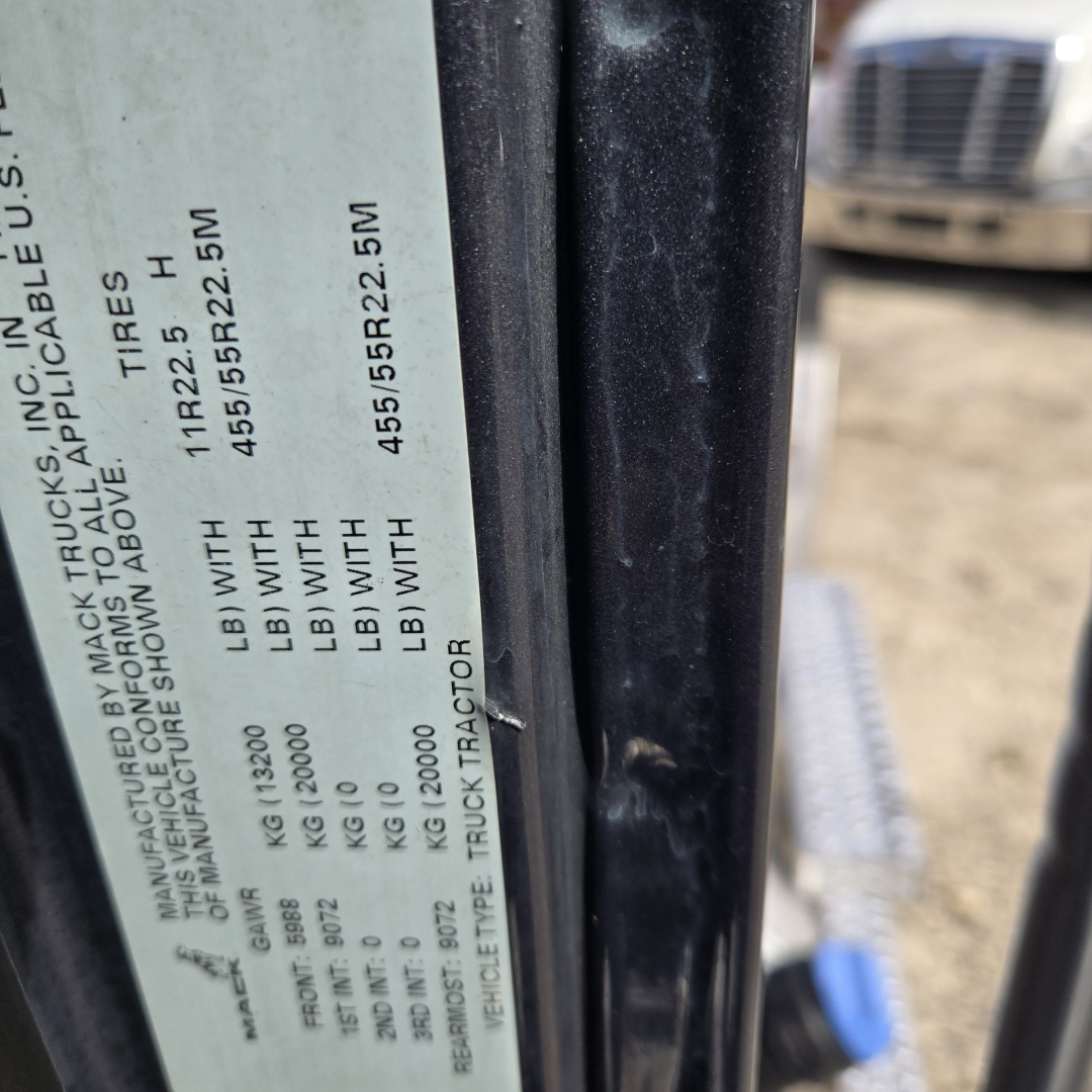 Label on Mack ANTHEN truck door showing tire sizes and axle weights with vertical text on a beige sticker; background shows another truck cab.