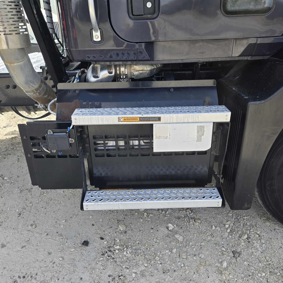 Truck side step area showing access stairs and toolbox mounted under cab; metal diamond-plate steps, black compartment, and small electronic box visible.