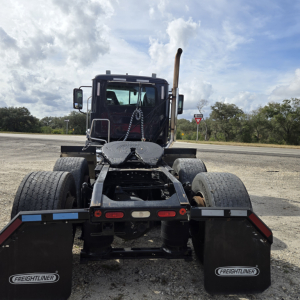 Truck chassis rear view showing dual rear axles, mudflaps with Freightliner logo, fifth wheel, and exhaust stack behind cab.