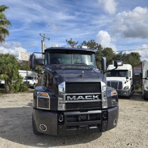 Front view of a dark blue Mack ANTHEM day cab truck showing large grille with MACK emblem, headlights, and bumper; other trucks in background.