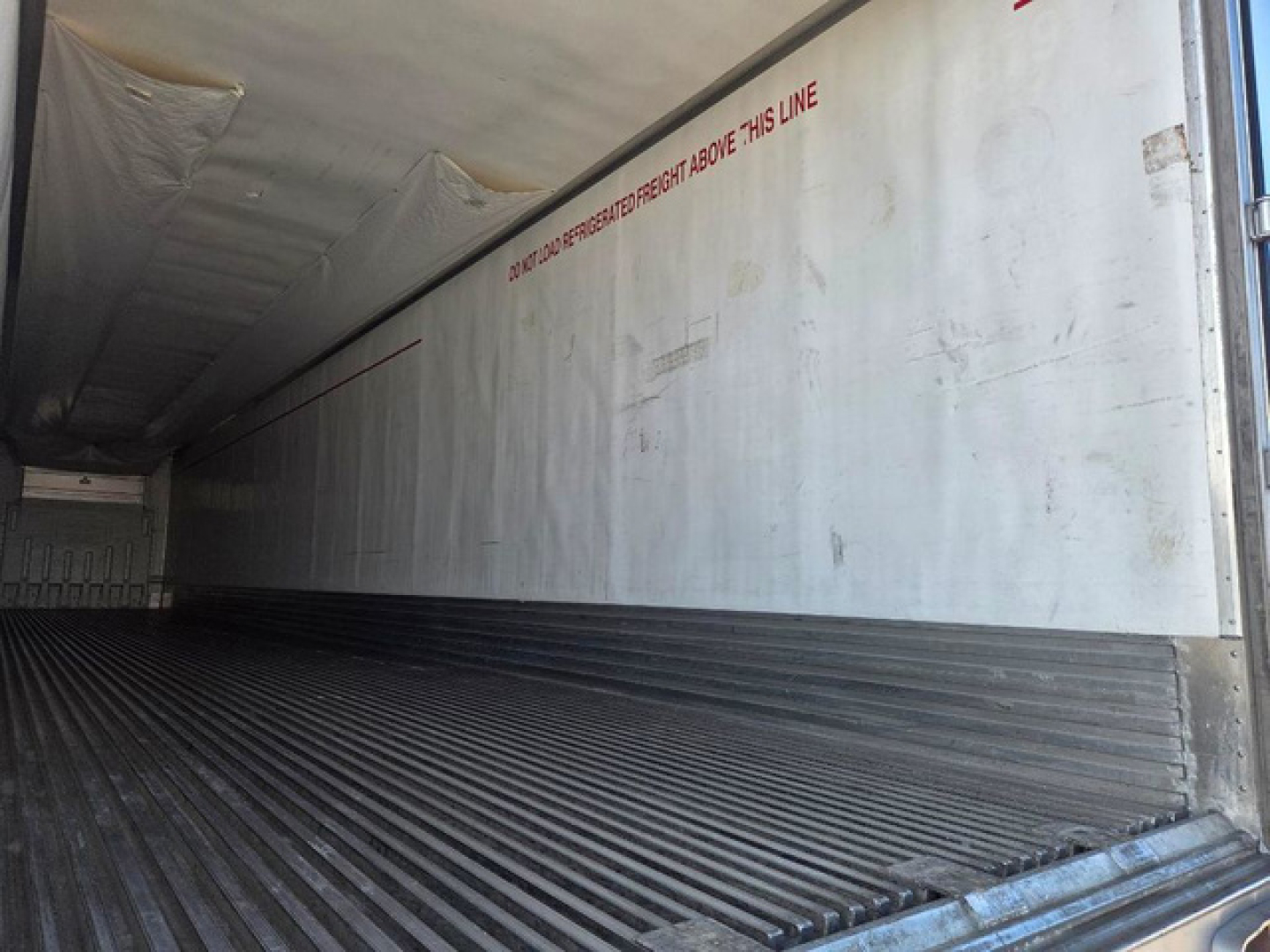 Inside of a refrigerated trailer with ribbed floor and white interior walls. Red text along wall reads: 'DO NOT OPEN THIS LINE' and other faded markings; trailer interior shows corrugated steel floor and light entry at rear.