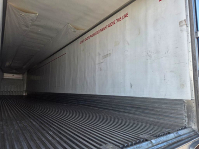 Inside of a refrigerated trailer with ribbed floor and white interior walls. Red text along wall reads: 'DO NOT OPEN THIS LINE' and other faded markings; trailer interior shows corrugated steel floor and light entry at rear.