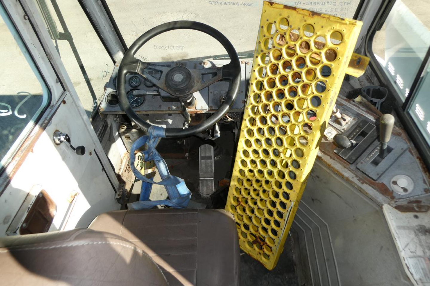 Truck cab interior with steering wheel and yellow perforated metal panel on the right; dashboard controls and blue seat belt visible.