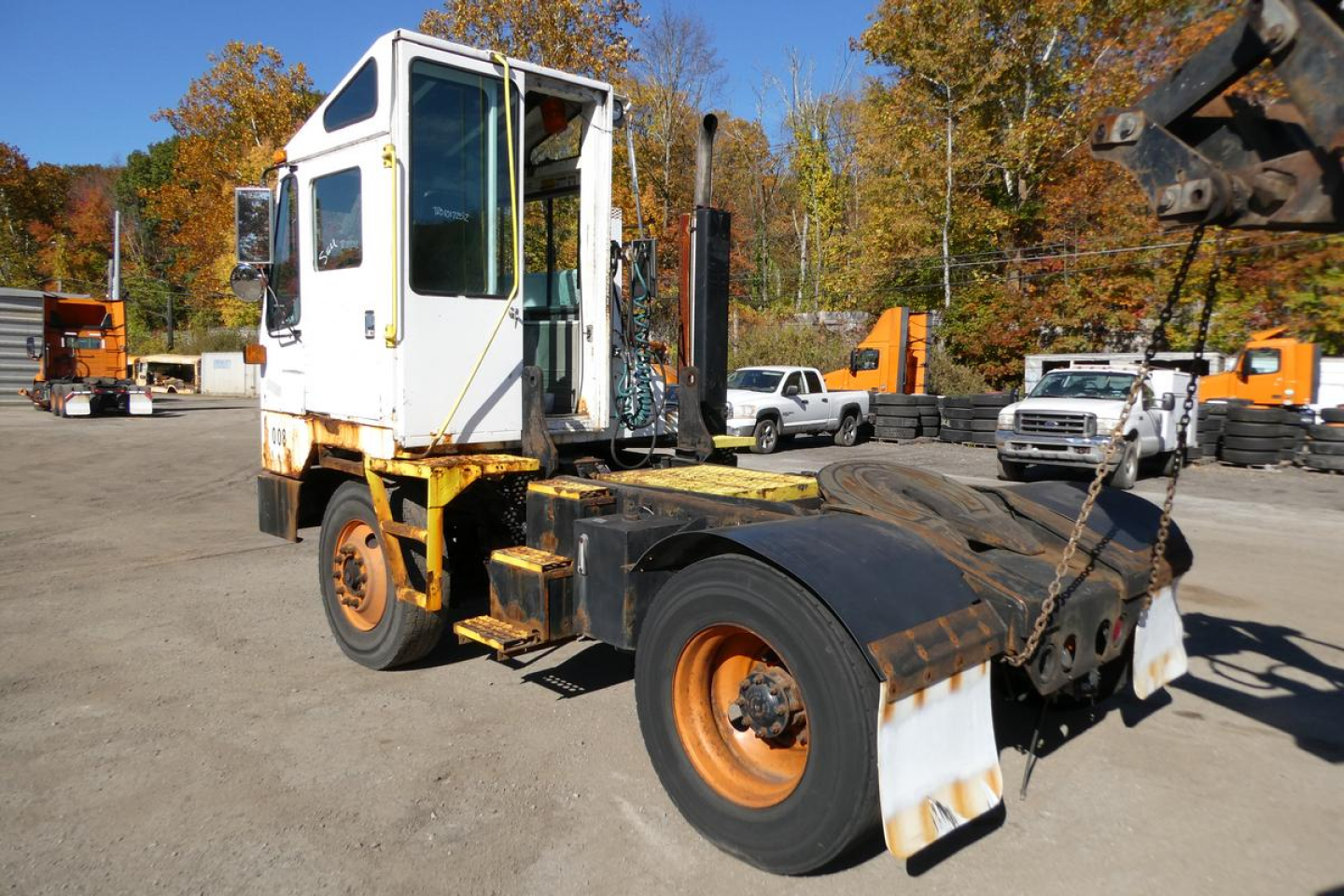 Truck tractor in yard with white cab and orange wheels, hooklift equipment, and fall foliage in background.