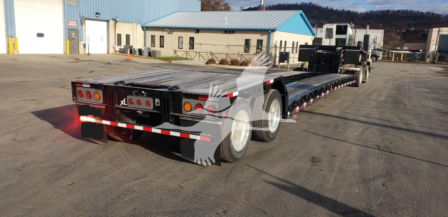 Trailer showing a long lowboy trailer with triple rear axles and reflective tape; rear lights and mud flaps visible.