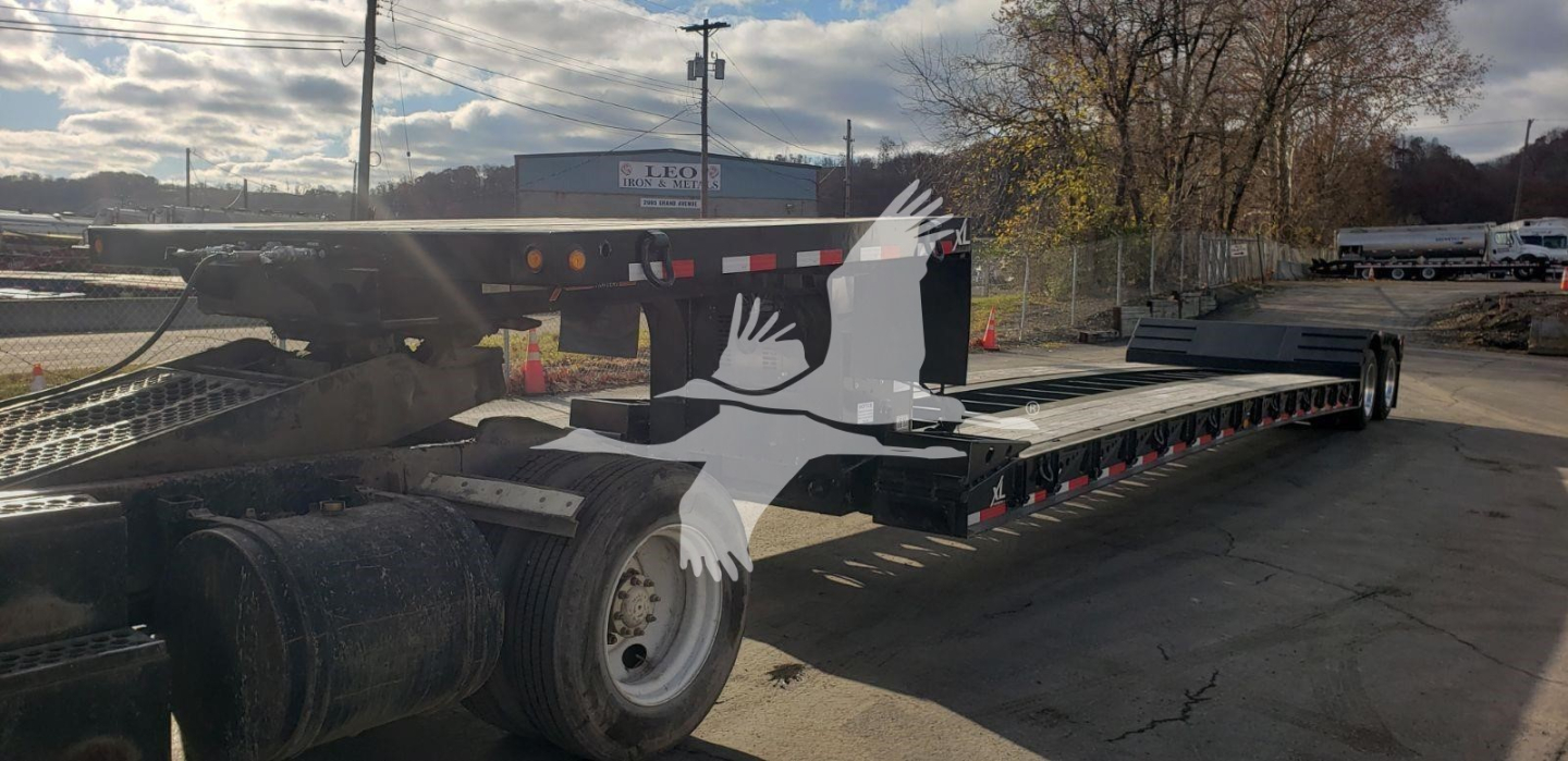 Lowboy trailer with long flat deck and multiple axles, side rails, and dual rear axles; visible decals and light reflectors along the side.
