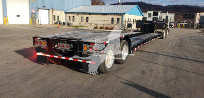 Trailer showing a long lowboy trailer with triple rear axles and reflective tape; rear lights and mud flaps visible.